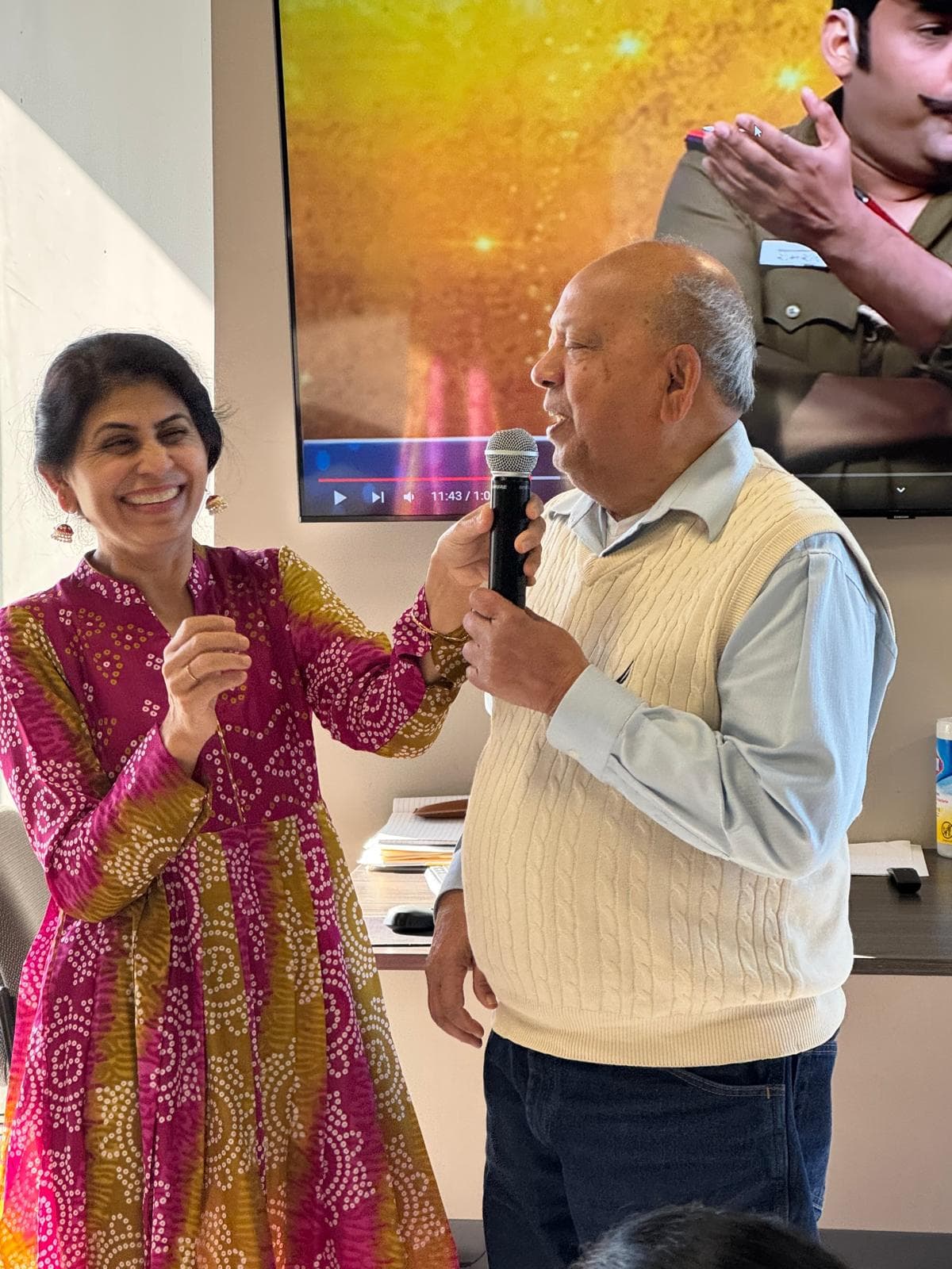 A member singing into a microphone while a staff member laughs beside him